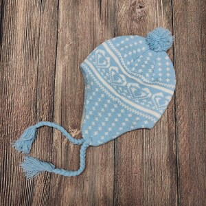 Puede incluir: Un gorro de punto azul claro con diseños blancos de copos de nieve y corazones. El gorro tiene orejeras, lazos trenzados con borlas y un pompón. Hecho de un material suave y cálido, ideal para el clima frío.