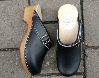 Puede incluir: Un par de zuecos de cuero negro con base de madera y una correa con hebilla. Los zuecos tienen un detalle de costura blanca alrededor del borde de la suela. La base de madera tiene una etiqueta que dice "Original Swedish Clogs".