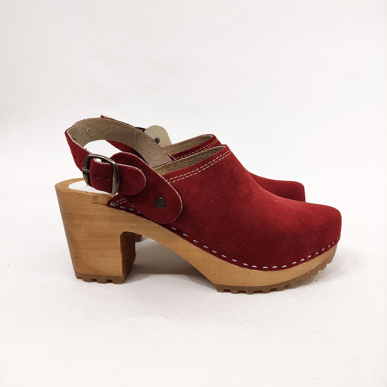 May include: Red suede clog with a wooden heel and a buckle strap. The clog has a white stitch detail around the sole.
