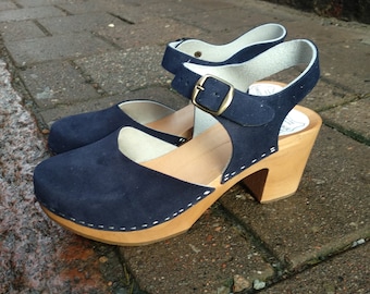 May include: Navy blue suede clogs with a wooden heel and a white stitch detail around the sole. The clogs have an ankle strap with a gold buckle.