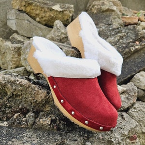 Op de afbeelding: Rode suède klompen met een witte imitatiebont voering en houten zolen. De klompen hebben decoratieve zilveren studs rond de randen.