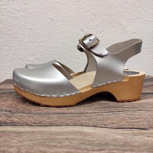 Puede incluir: Sandalias de madera con tacón de madera y correa de hebilla en cuero plateado. Las sandalias están sobre una superficie de madera.