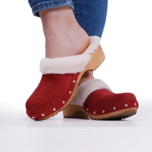 Op de afbeelding: Rode suède klompen met een witte nep bontkraag en een houten hak. De klompen hebben een studded ontwerp aan de voorkant en een rubberen zool.