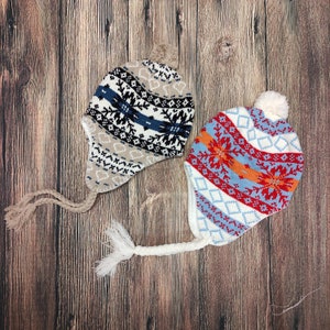 Puede incluir: Dos gorros de invierno de punto con orejeras y pompones. Un gorro presenta un patrón beige, negro, blanco y azul. El otro gorro tiene un diseño rojo, naranja, azul y blanco. Ambos gorros tienen lazos trenzados.