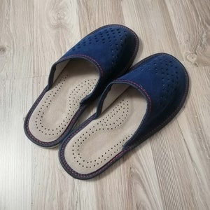 Puede incluir: Un par de zapatillas de ante azul marino con un dise&ntilde;o perforado en la parte superior. Las zapatillas tienen un interior beige claro con un patr&oacute;n punteado y costuras rojas en los bordes. Sobre una superficie de madera marr&oacute;n claro.