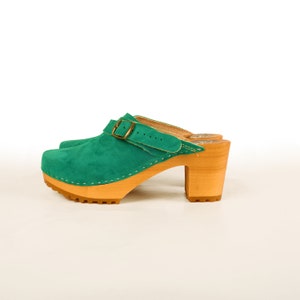 May include: Green suede heeled clogs with a buckle closure and a wooden platform sole.
