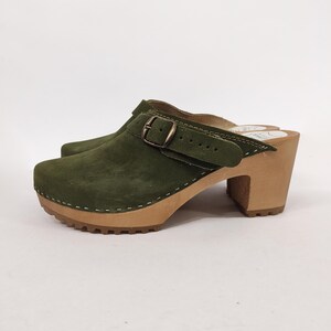 May include: Green suede clog with a buckle and a wooden heel. The clog has a stitched seam around the edge of the sole.