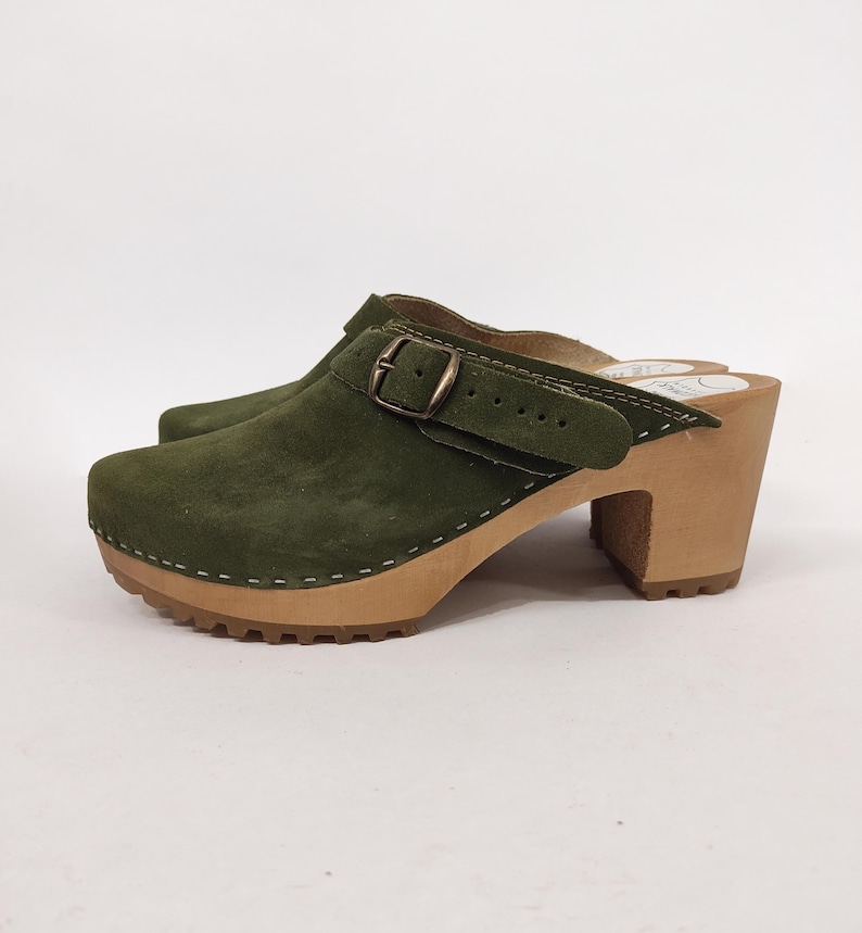 May include: Green suede clog with a buckle and a wooden heel. The clog has a stitched seam around the edge of the sole.