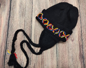 May include: A black knit hat with ear flaps and a tassel. The hat features a colorful embroidered design in red, yellow, and blue. The hat has long braided ties with tassels at the ends.