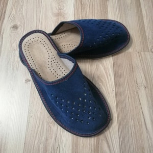Puede incluir: Un par de zapatillas de ante azul marino con un dise&ntilde;o perforado en la parte superior. Las zapatillas tienen un ribete rojo y un interior marr&oacute;n claro. Dise&ntilde;adas para uso en interiores, est&aacute;n sobre una superficie de madera.