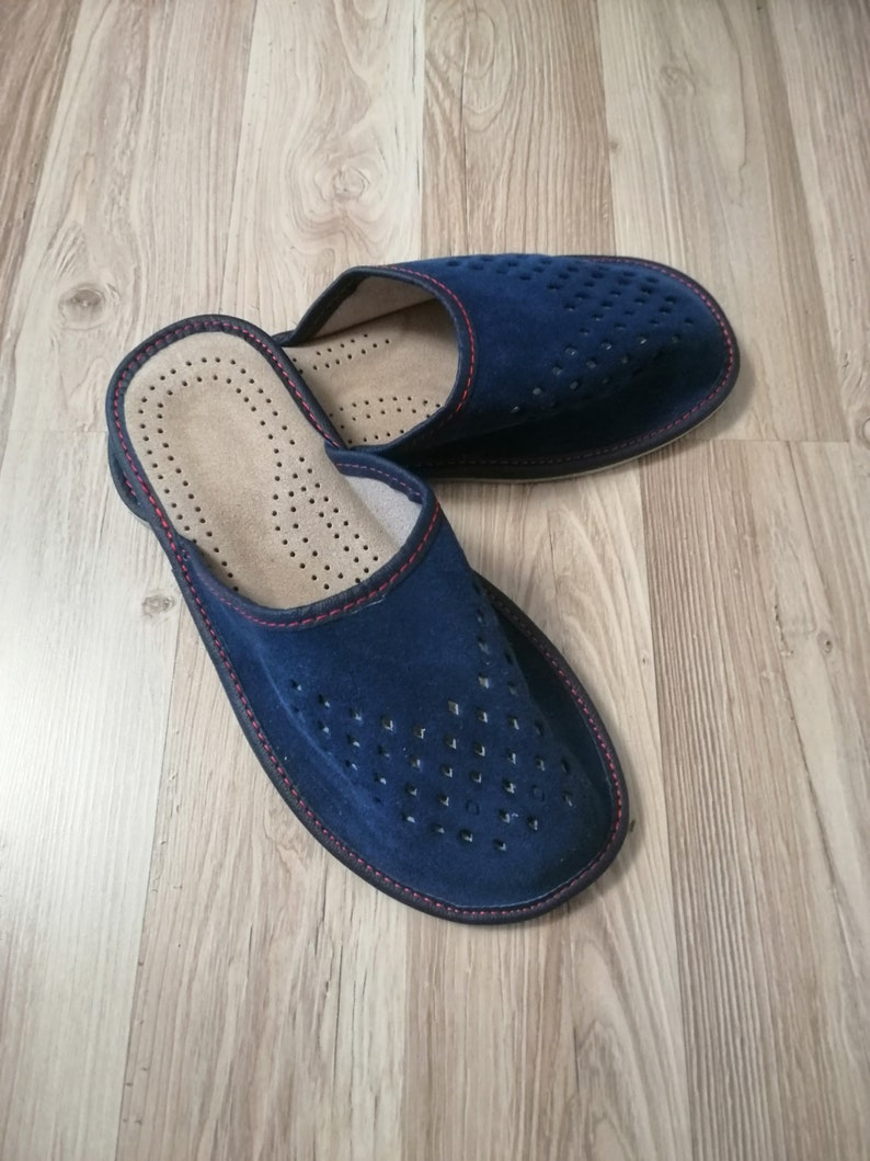 Puede incluir: Un par de zapatillas de ante azul marino con un dise&ntilde;o perforado en la parte superior. Las zapatillas tienen un ribete rojo y un interior marr&oacute;n claro. Dise&ntilde;adas para uso en interiores, est&aacute;n sobre una superficie de madera.