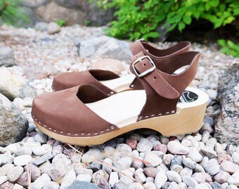 May include: Brown leather clogs with an ankle strap and buckle. The clogs have a wooden sole and are sitting on a bed of small white stones.