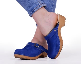 May include: A pair of blue suede clogs with a buckle strap and wooden soles. The clogs are worn by a person wearing blue jeans.