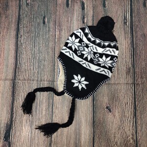 Puede incluir: Gorro de invierno de punto negro y blanco con un pompón en la parte superior y un patrón de copo de nieve. El gorro tiene orejeras y un largo borlón.