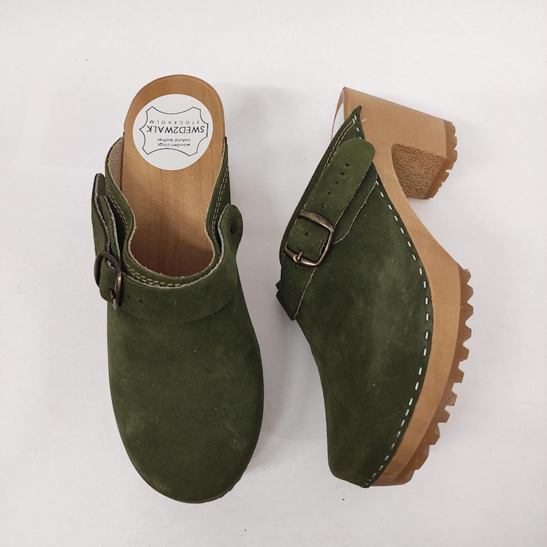 May include: A pair of green suede clogs with a buckle strap and a wooden platform sole. The clogs are made by the brand "Swedish Walk" and are made in Stockholm.