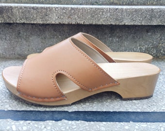 May include: A pair of brown leather clogs with a wooden heel. The clogs have a simple design with a single strap across the top of the foot.