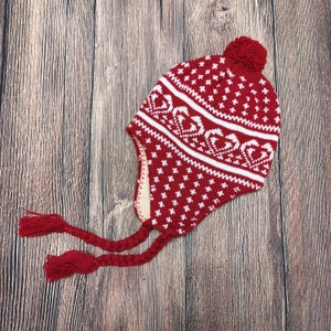 Puede incluir: Gorro de punto rojo y blanco con un diseño de corazón y estrella, orejeras y borlas trenzadas. El gorro tiene un pompón rojo en la parte superior. El gorro está hecho de un material suave y cálido.