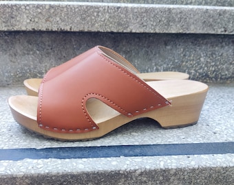 May include: A pair of brown leather clogs with a wooden heel. The clogs have a cut-out design on the top of the foot.