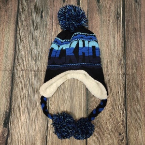 Puede incluir: Un gorro de invierno de punto azul marino con orejeras y un gran pompón. El gorro presenta la palabra "AKITA" en tonos azules y un forro polar blanco. Cintas trenzadas con pompones cuelgan de las orejeras.
