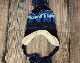 May include: A navy blue knit winter hat with ear flaps and a large pom-pom. The hat features the word "AKITA" in shades of blue and a white fleece lining. Braided ties with pom-poms hang from the ear flaps.