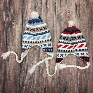 Puede incluir: Dos gorros de invierno con orejeras y borlas blancas. Los sombreros tienen un diseño estampado en azul, blanco, marrón y gris. Un sombrero tiene un estampado rojo y gris. Ambos sombreros tienen un pompón blanco en la parte superior.