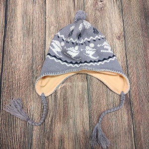 Puede incluir: Un gorro de invierno de punto gris con un patrón blanco y azul marino, orejeras y una borla. El gorro tiene un forro de color melocotón claro y un pompón gris en la parte superior. Las orejeras tienen lazos trenzados con borlas.