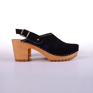 May include: Black suede heeled clog with a wooden platform and a slingback strap with a buckle. The clog has a white stitch detail around the edge of the platform.