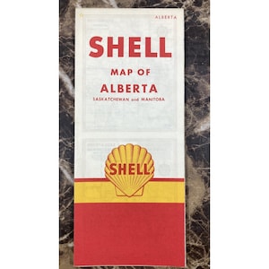 May include: A vintage Shell road map of Alberta, Saskatchewan, and Manitoba. The cover is white with red text reading SHELL MAP OF ALBERTA SASKATCHEWAN and MANITOBA. It features a yellow and red Shell scallop logo centered above a red horizontal band.