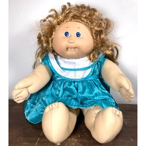 May include: A vintage Cabbage Patch Kids doll with blonde curly hair and blue eyes. The doll is wearing a teal dress with a white collar. The doll is sitting on a wooden surface.