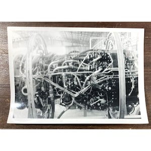 May include: A black and white photo of a complex engine with many pipes and wires. The engine is in a large, industrial setting.