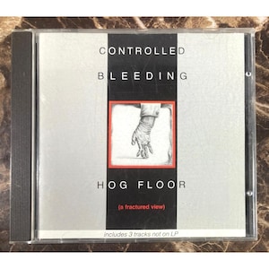 May include: A CD case with the text "CONTROLLED BLEEDING" and "HOG FLOOR" in black. The cover features a drawing of a gloved hand within a red-bordered square. The text "(a fractured view)" and "includes 3 tracks not on LP" are also present.
