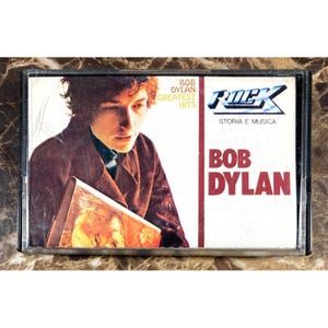 May include: A vintage cassette tape featuring Bob Dylan's Greatest Hits album. The cover art shows a portrait of Bob Dylan in a brown jacket. The cassette case is black, and the album title is in red.