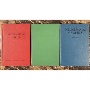 May include: Three vintage hardcover books are displayed. The books are in red, green, and blue. The red book is titled "MATHEMATICAL TABLES". The green book has a blank cover. The blue book is titled "COLLEGE MANUAL OF OPTICS".