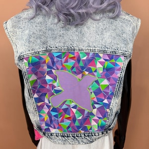 May include: A sleeveless acid-wash denim vest featuring a large rectangular back panel with a vibrant geometric pattern of purple, blue, pink, and green triangles. A solid light purple bird silhouette is appliqued in the center.