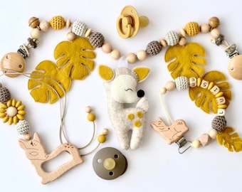 May include: A yellow and brown baby teether with a wooden deer, a sunflower, and a felt monstera leaf. The teether also includes a wooden bird, a pacifier, and a name tag with the letters "Elise".