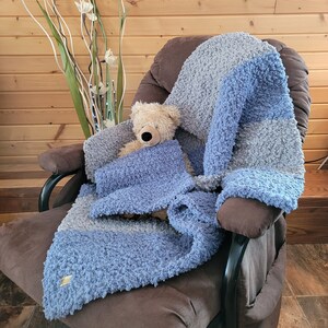 May include: A plush, blue and gray striped throw blanket is draped over a brown armchair. A teddy bear is sitting on the blanket.