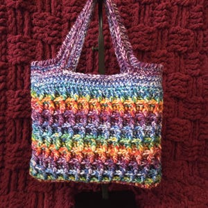 May include: A rectangular crochet tote bag featuring a vibrant, multi-colored variegated yarn pattern with hues of blue, purple, green, yellow, and orange. The bag has a textured waffle-like stitch design and two matching crochet handles.