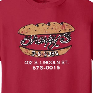 May include: Red t-shirt with a vintage-style logo for "Sharpy's 3-S-Subs" featuring a sandwich graphic and the text "402 S. LINCOLN ST. 678-0015."