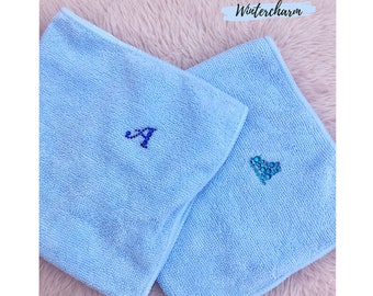 Peut inclure: Deux serviettes en microfibre bleu clair avec des motifs brodés. Une serviette a une lettre "A" en strass bleu et l'autre a un cœur en strass bleu. Les serviettes sont sur une surface rose duveteuse. Wintercharm est écrit en blanc sur fond noir.