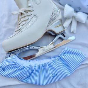 Op de afbeelding: Ivoor schaatsen met zilveren messen en strass accenten. De schaatsen liggen op een wit oppervlak met een blauw-wit gestreepte schaatsbeschermer en een bijpassende strik.