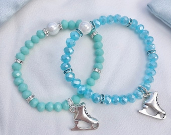 May include: Two light blue beaded bracelets with silver ice skate charms. The bracelets have small white pearl accents and are displayed on a white surface.
