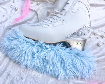 May include: White figure skate with a blue fluffy boot cover. The skate has a gold trim and is decorated with rhinestones. The skate is sitting on a white fluffy surface with blue and pink feathers.