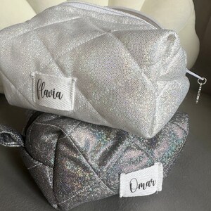 Può includere: Due trousse da toilette in glitter argento con etichette bianche. La trousse superiore ha il nome "flavia" sull'etichetta. La trousse inferiore ha il nome "Omar" sull'etichetta.