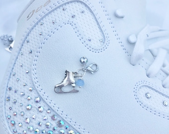 Peut inclure: Botte de patinage artistique blanche avec un charme argenté en forme de patins à glace, un cristal bleu et un cœur argenté. La botte est décorée de strass irisés et porte l'inscription "Goo-Fly" sur le côté.