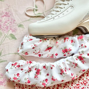 May include: White figure skates with a floral print skate guard. The skate guard is white with a red floral pattern.