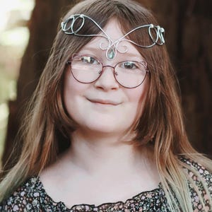 May include: A silver-toned wire circlet with a central green gemstone and delicate swirls. The circlet is worn on the head. The person is wearing cat-eye glasses with a red frame and a floral print top.