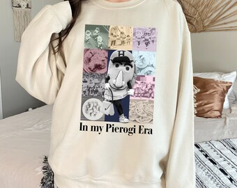 May include: A cream-colored sweatshirt with a collage of images featuring a cartoon pierogi mascot wearing a baseball uniform and holding a bat. The text "In my Pierogi Era" is printed below the collage.