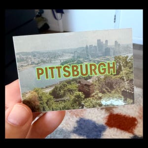 May include: A rectangular postcard featuring a scenic view of Pittsburgh, Pennsylvania. The image showcases the city skyline, bridges, and river, with the word "PITTSBURGH" in green and brown block letters across the top.