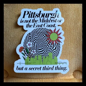 May include: A white sticker with black text that reads "Pittsburgh is not the Midwest or the East Coast, but a secret third thing." The sticker features a black and white swirling design with a green sun and green city skyline.