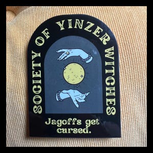 May include: Black and yellow sticker with an arch design. The text "Society of Yinzer Witches" is written in a circle around the arch. Inside the arch, two hands are shown with a yellow moon in between them. The text "Jagoffs get cursed." is written below the arch.